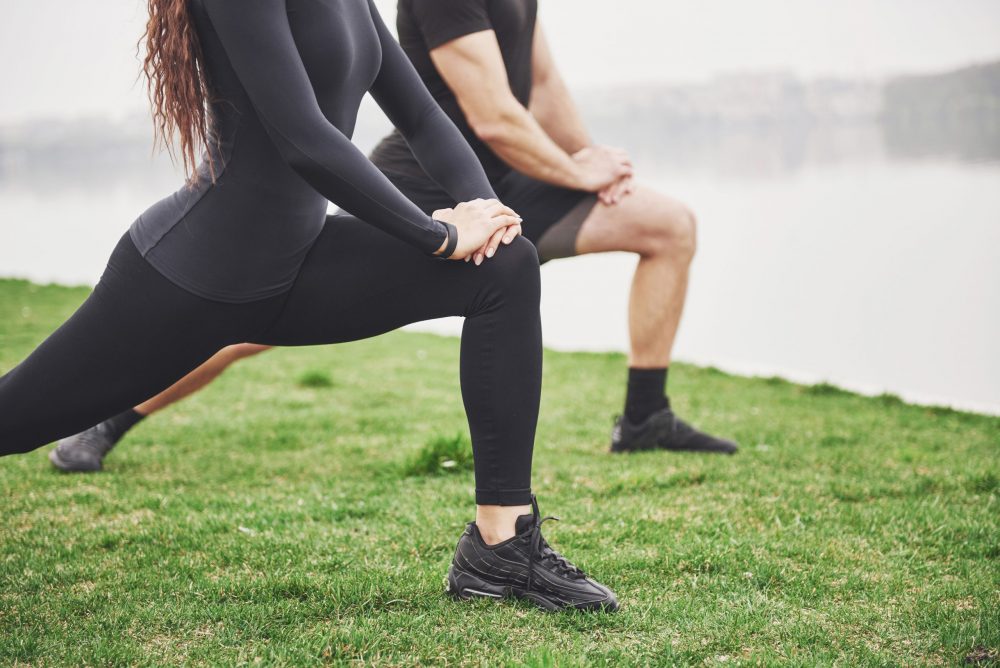 Fitness couple stretching outdoors in park near the water. Young bearded man and woman exercising together in morning